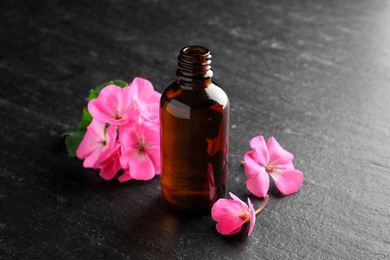 Photo of Bottle of geranium essential oil and beautiful flowers on black table, closeup