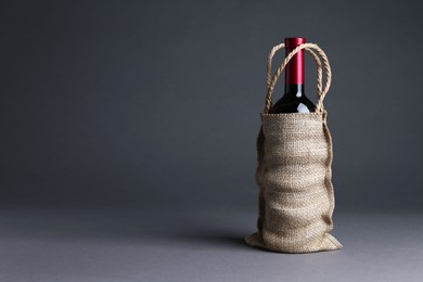 Photo of Wine bottle in burlap bag on grey background, space for text