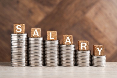 Photo of Stacked coins and cubes with word Salary on light wooden table, closeup