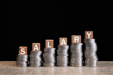 Photo of Stacked coins and cubes with word Salary on wooden table against black background, closeup