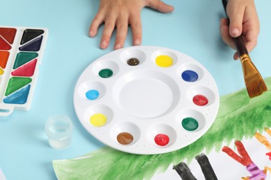 Photo of Boy with brush, palette, paints and picture at light blue table, closeup