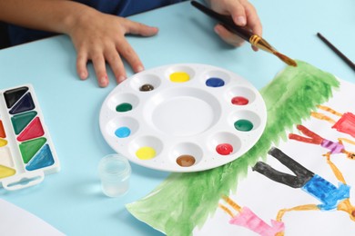 Boy with brush, palette, paints and picture at light blue table, closeup