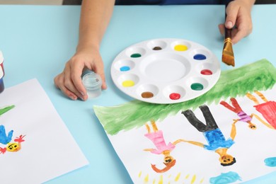 Photo of Boy with brush, container of water, palette, paints and picture at light blue table, closeup