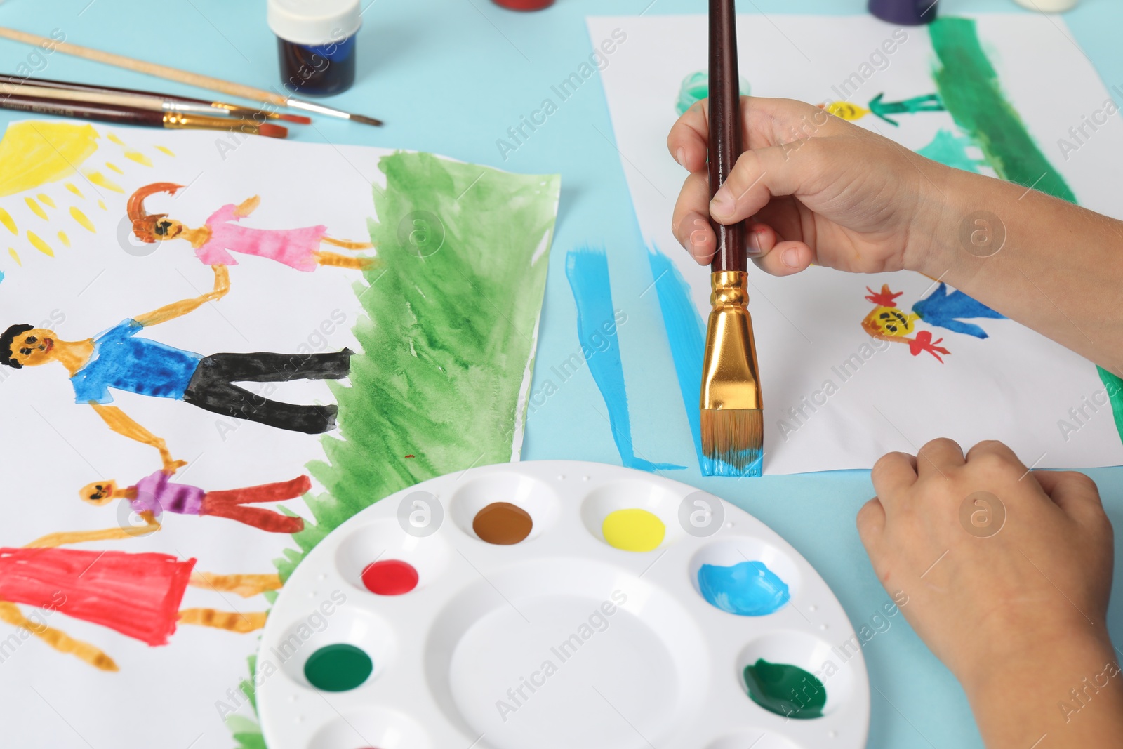 Photo of Boy drawing his family with paint at light blue table, closeup