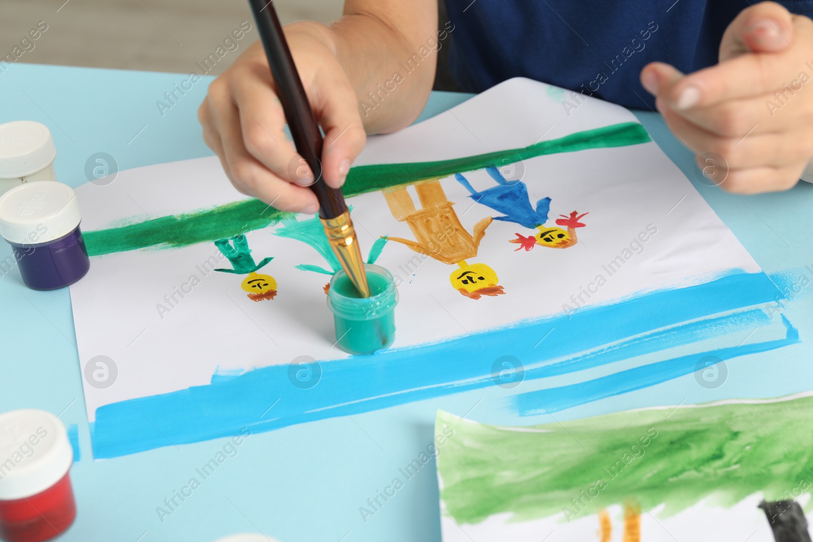 Photo of Boy drawing his family with paint at light blue table, closeup