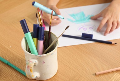 Cup with felt pens, brushes and boy drawing picture at wooden table, selective focus