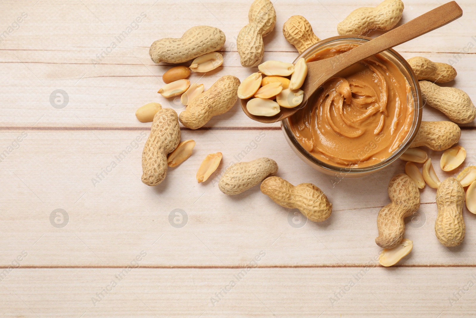 Photo of Tasty peanut butter in bowl and spoon with groundnuts on wooden table, flat lay. Space for text