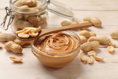 Tasty peanut butter in bowl, groundnuts, jar and spoon on wooden table, closeup