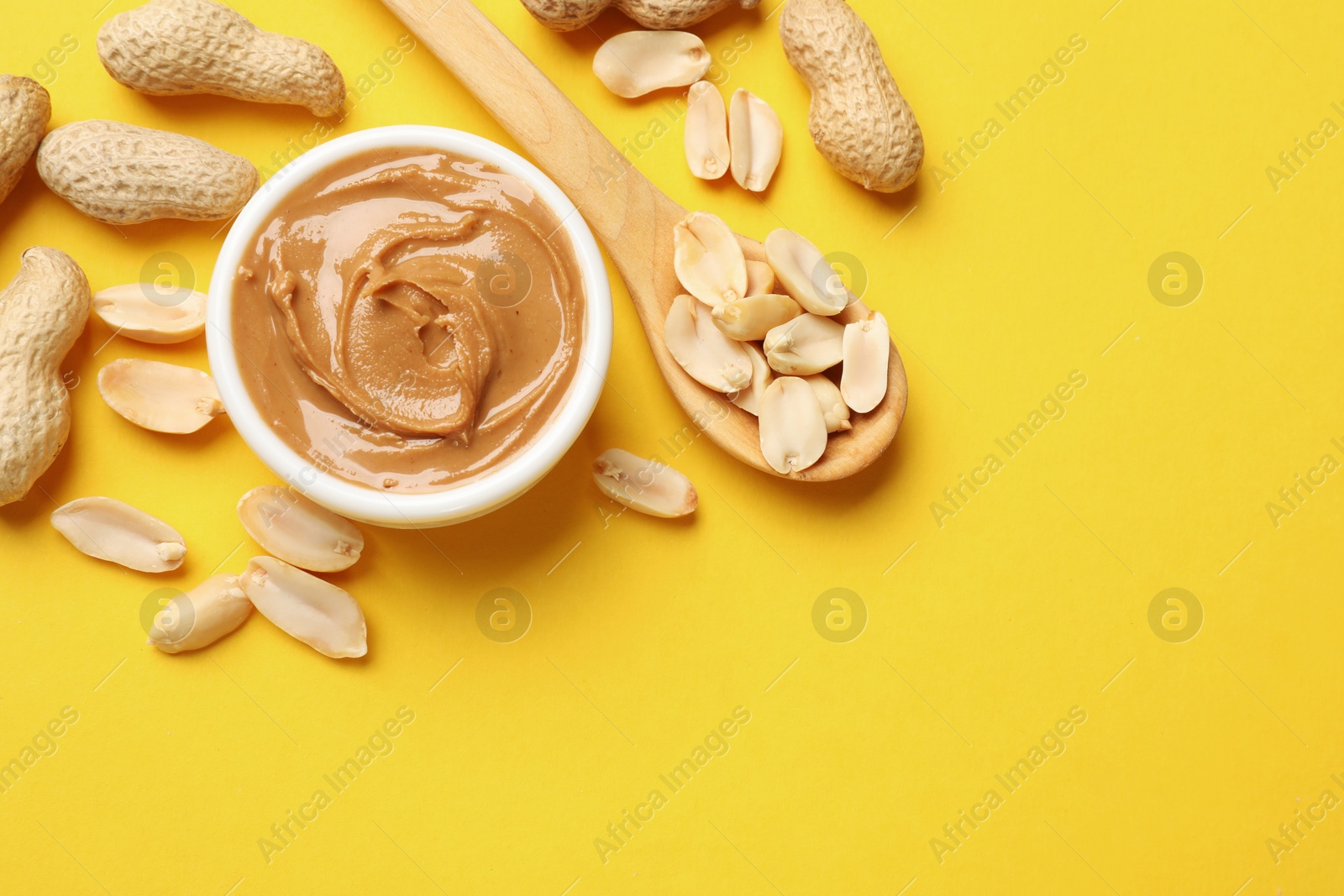 Photo of Tasty peanut butter in bowl and spoon with groundnuts on yellow table, flat lay. Space for text