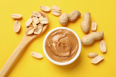 Photo of Tasty peanut butter in bowl and spoon with groundnuts on yellow table, flat lay