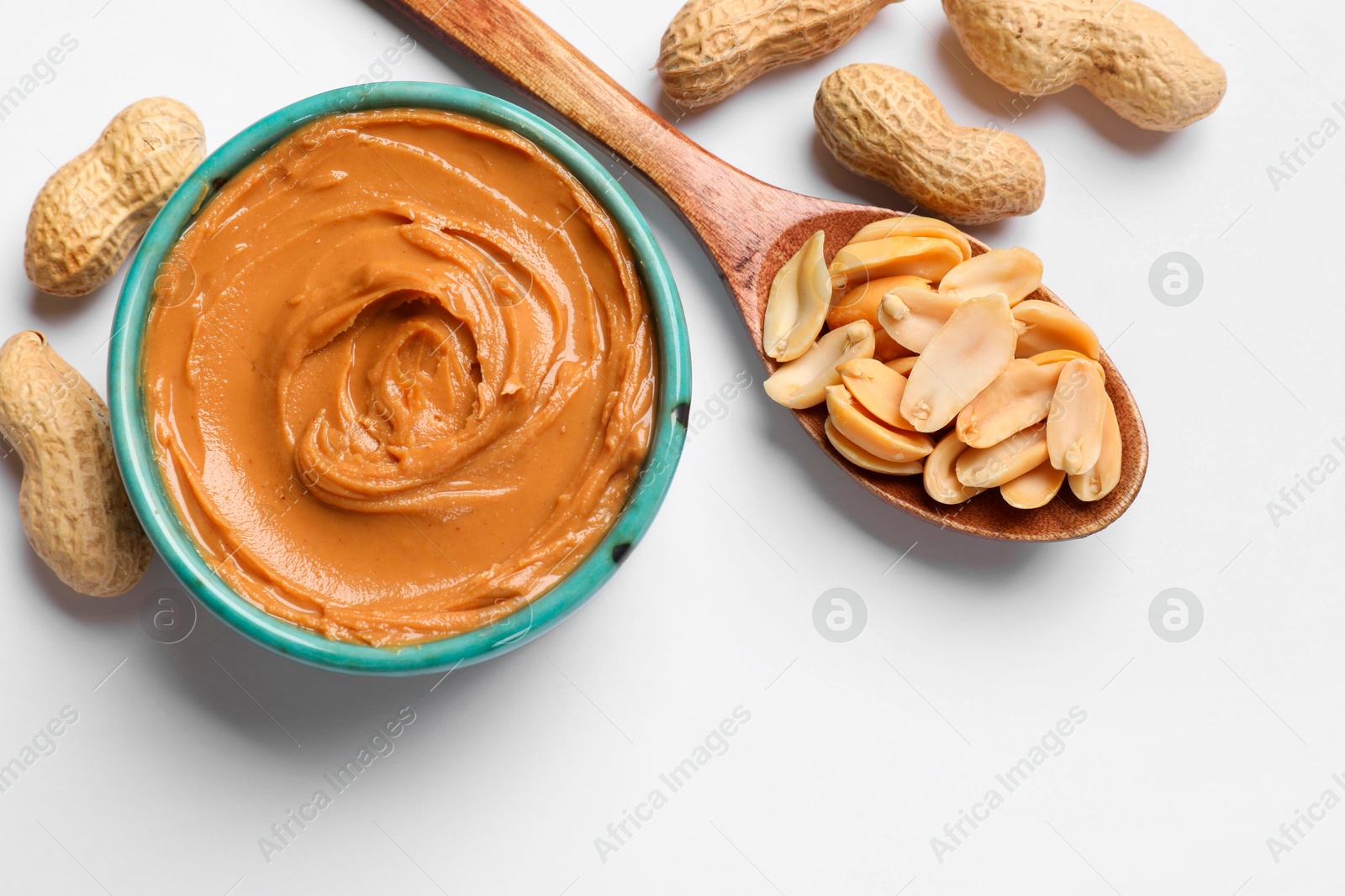 Photo of Tasty peanut butter in bowl and spoon with groundnuts on white table, flat lay