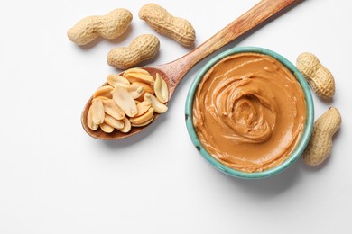 Photo of Tasty peanut butter in bowl and spoon with groundnuts on white table, flat lay