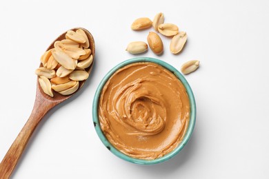 Photo of Tasty peanut butter in bowl and spoon with groundnuts on white table, flat lay