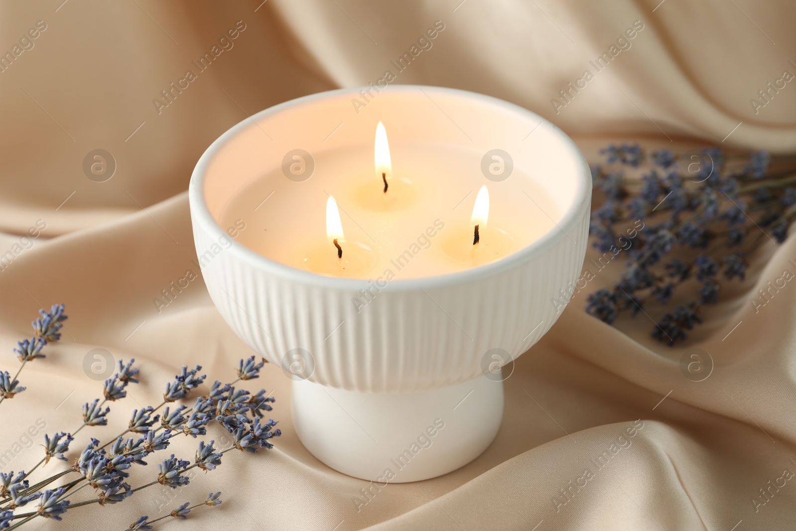 Photo of Burning candle and flowers on beige fabric, closeup