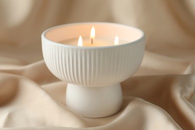 Stylish burning candle on beige fabric, closeup