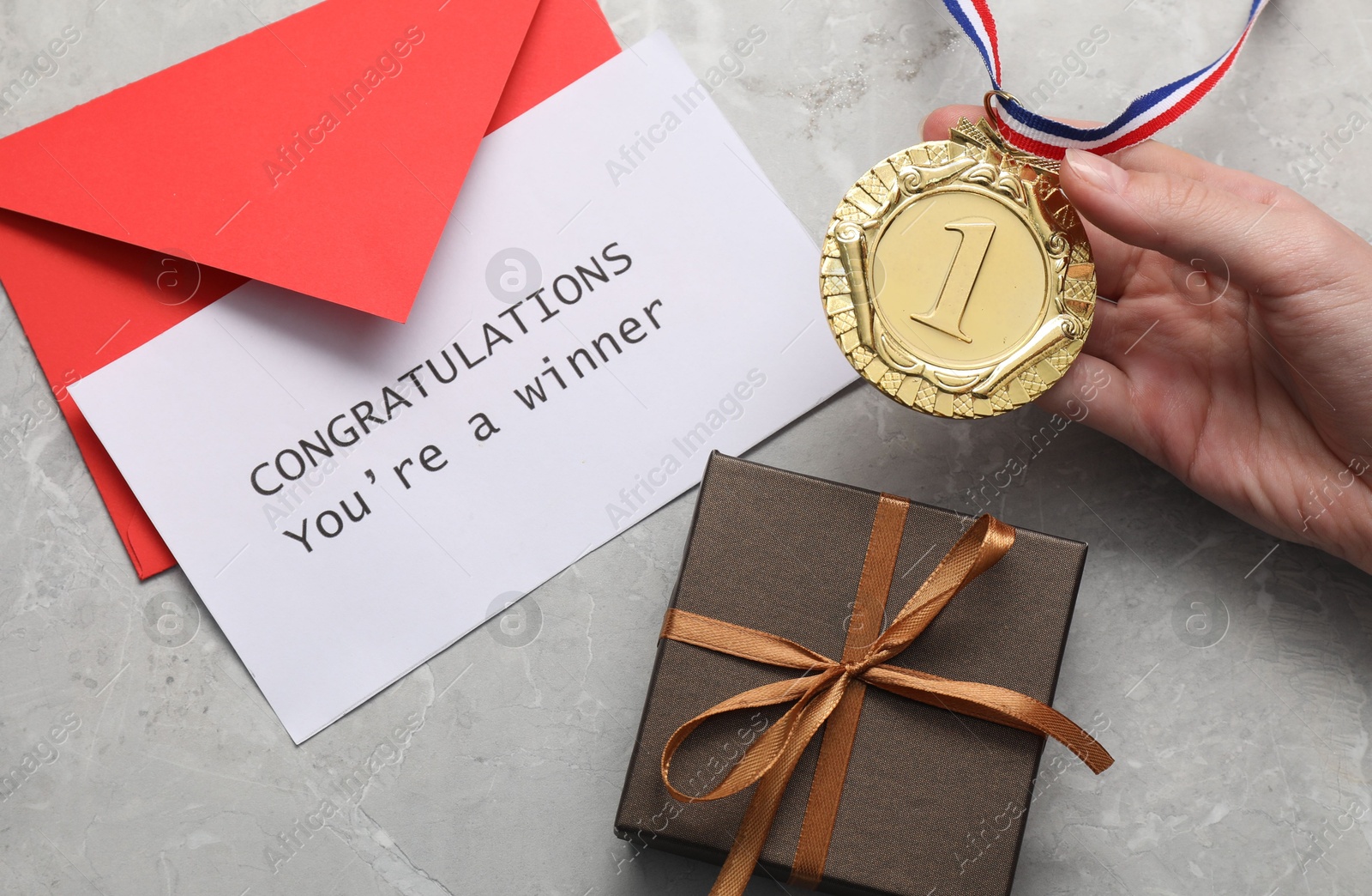 Photo of Woman holding golden medal, card with phrase Congratulations You`re Winner, gift box and envelope at grey marble table, top view