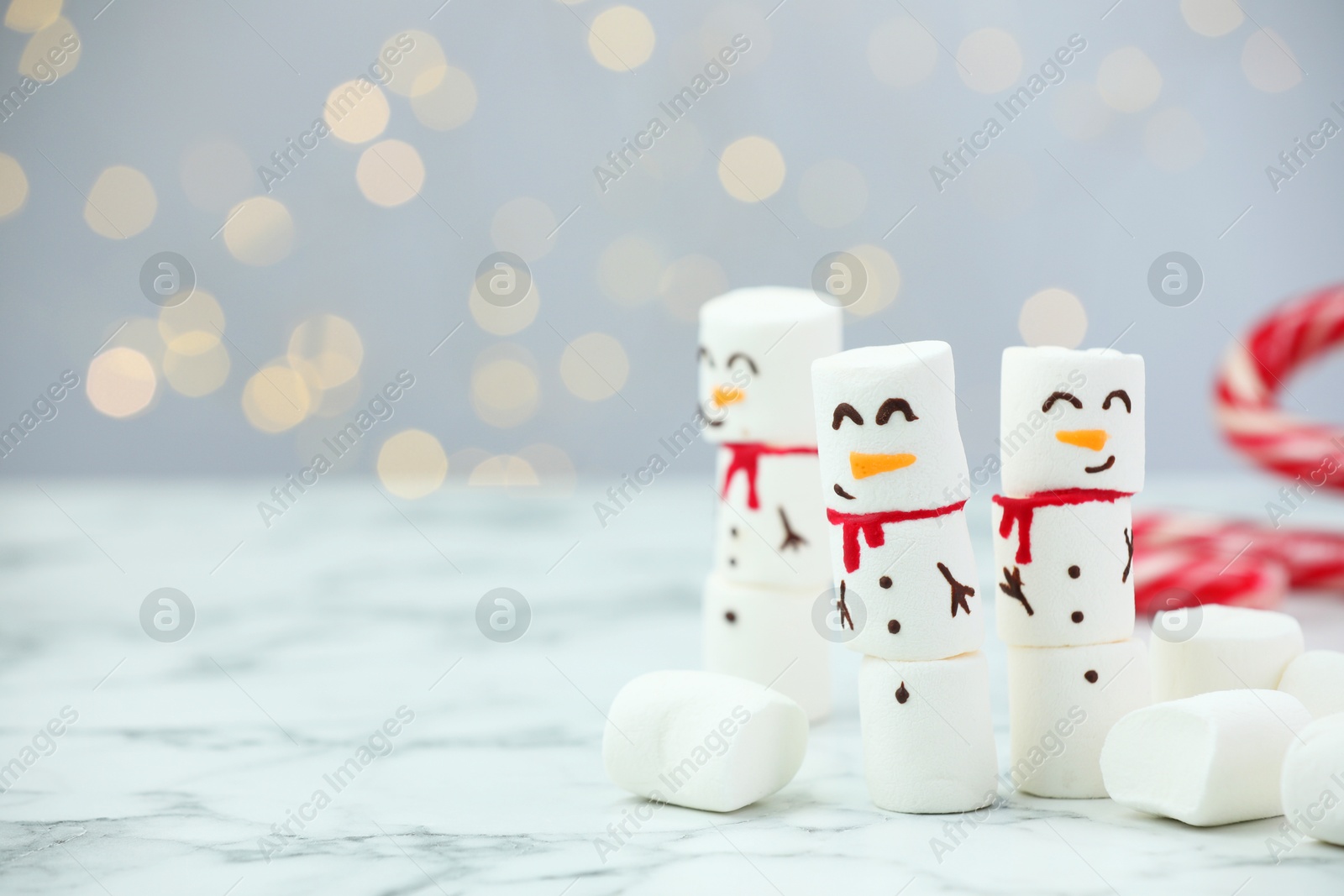 Photo of Funny snowmen made with marshmallows on white table against blurred lights. Space for text