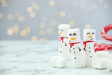 Photo of Funny snowmen made with marshmallows on white table against blurred lights. Space for text