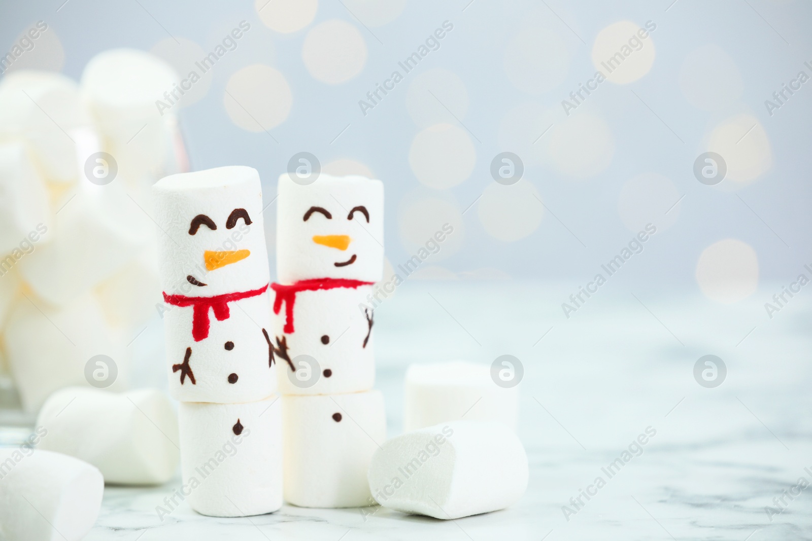 Photo of Funny snowmen made with marshmallows on white table against blurred lights. Space for text