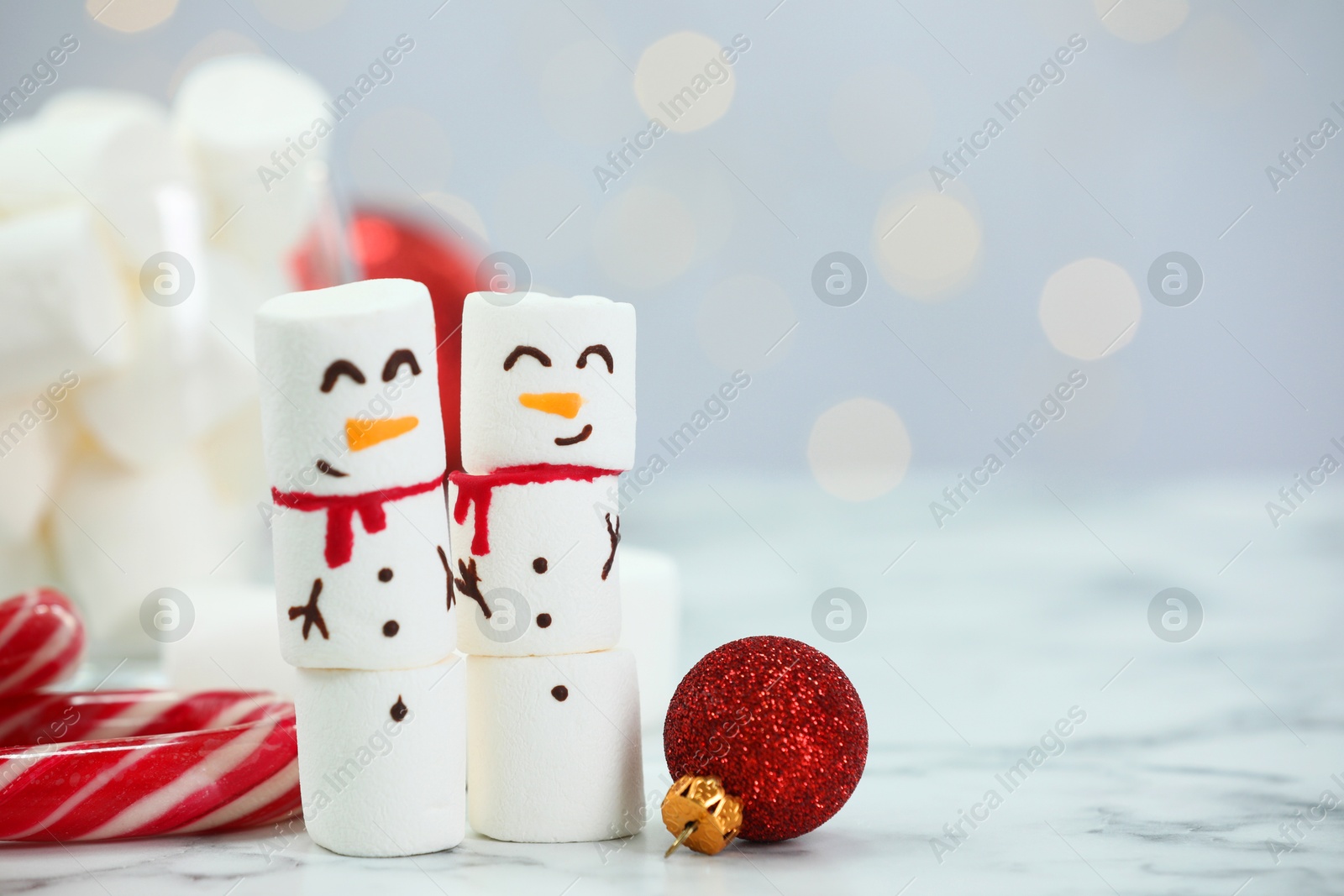 Photo of Funny snowmen made with marshmallows and Christmas decor on white table against blurred lights, closeup. Space for text