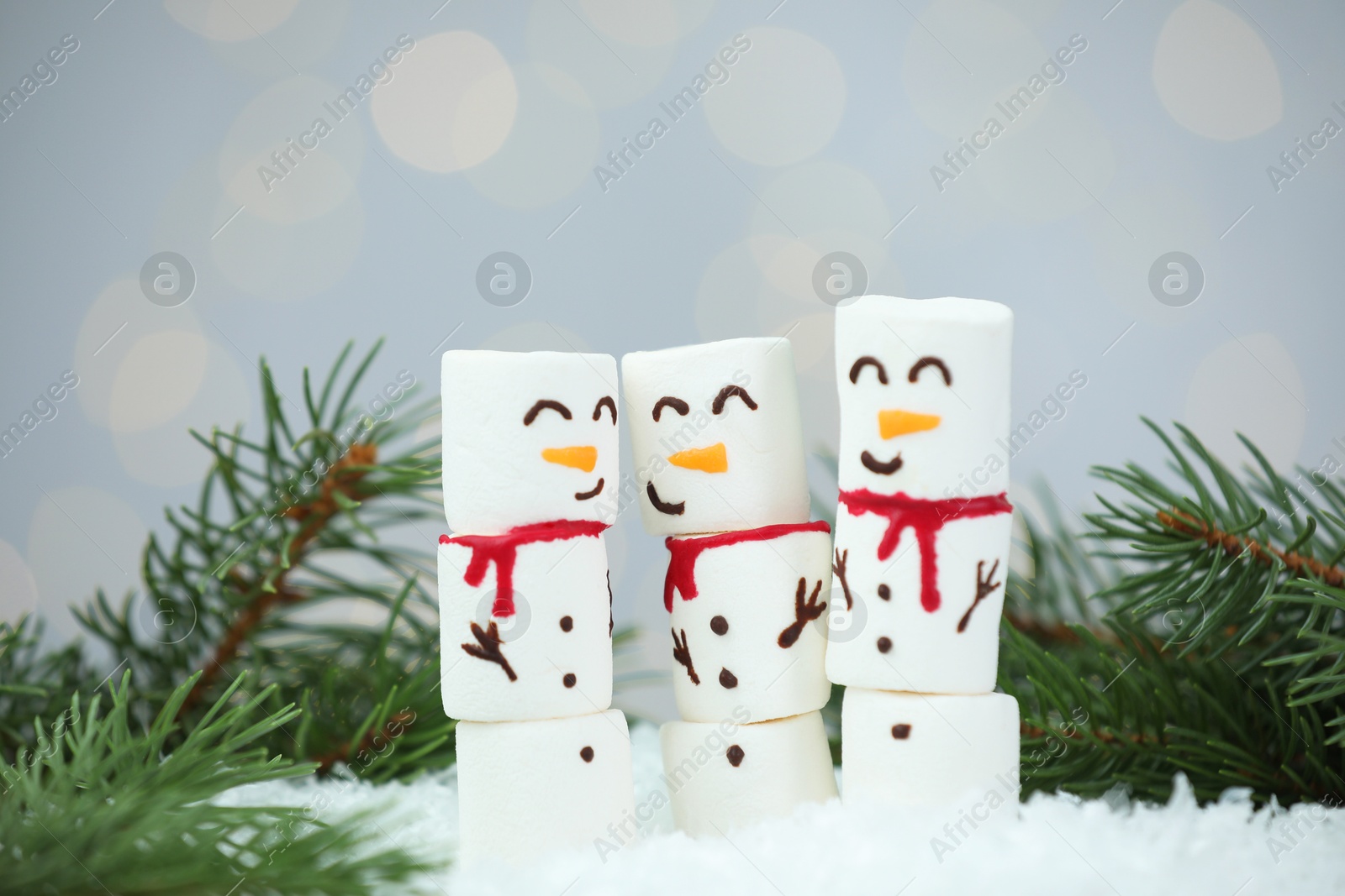 Photo of Funny snowmen made with marshmallows and fir tree branches on artificial snow against blurred lights, closeup