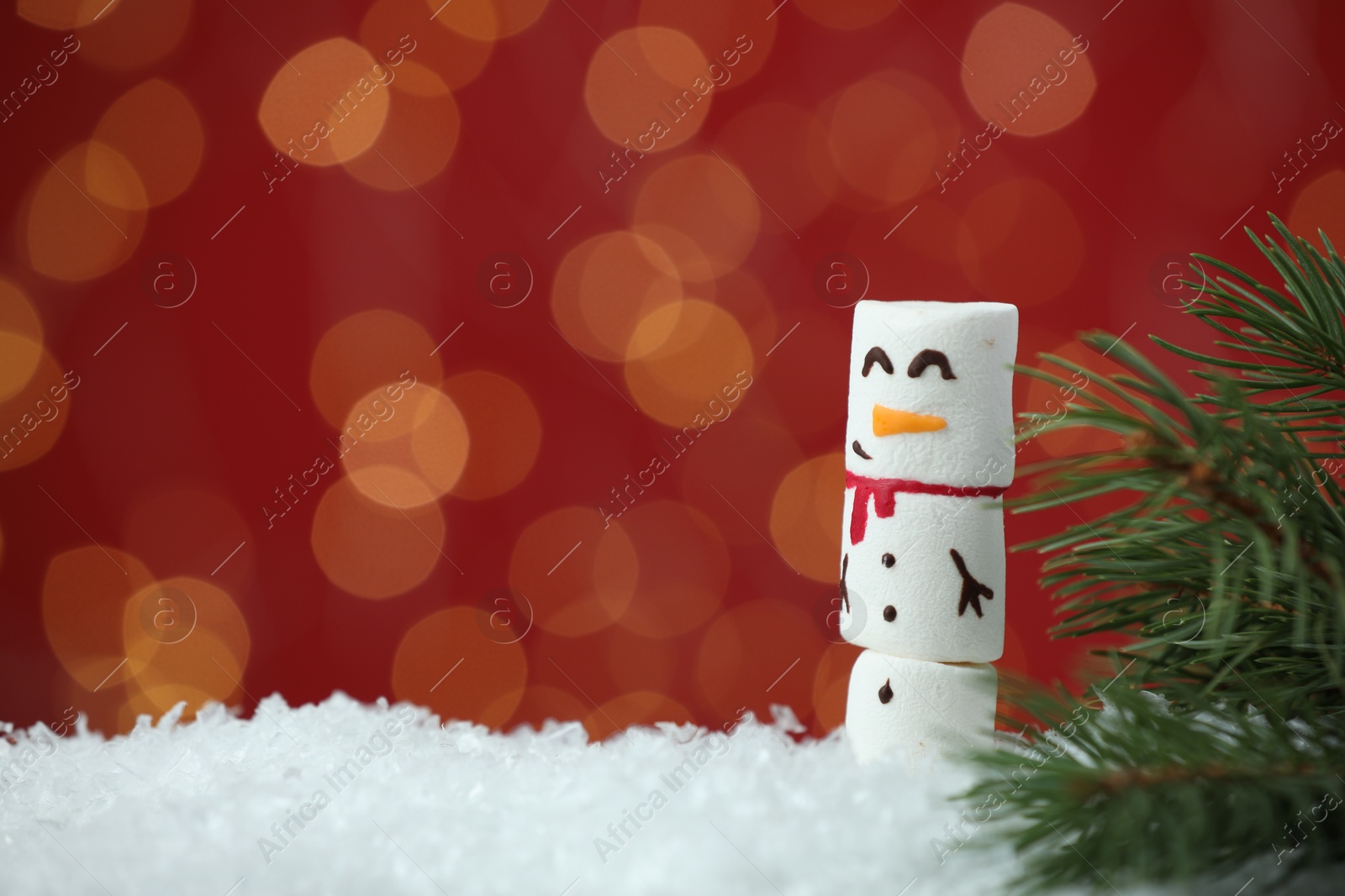 Photo of Funny snowman made with marshmallows and fir tree branch on artificial snow against blurred festive lights. Space for text