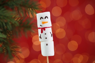 Photo of Funny snowman made of marshmallows on red background with fir tree branch and blurred lights