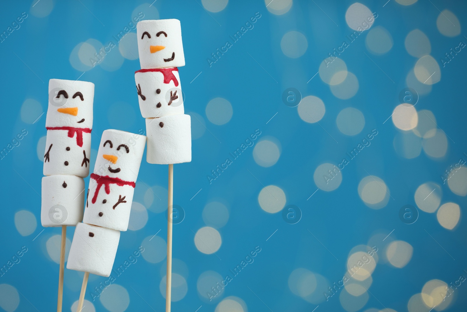 Photo of Funny snowmen made of marshmallows on blue background with blurred lights. Space for text