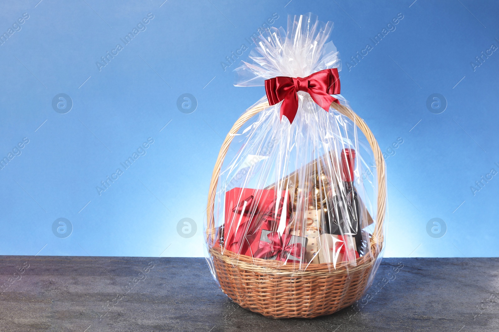 Photo of Wicker gift basket with wine on grey textured table against light blue background