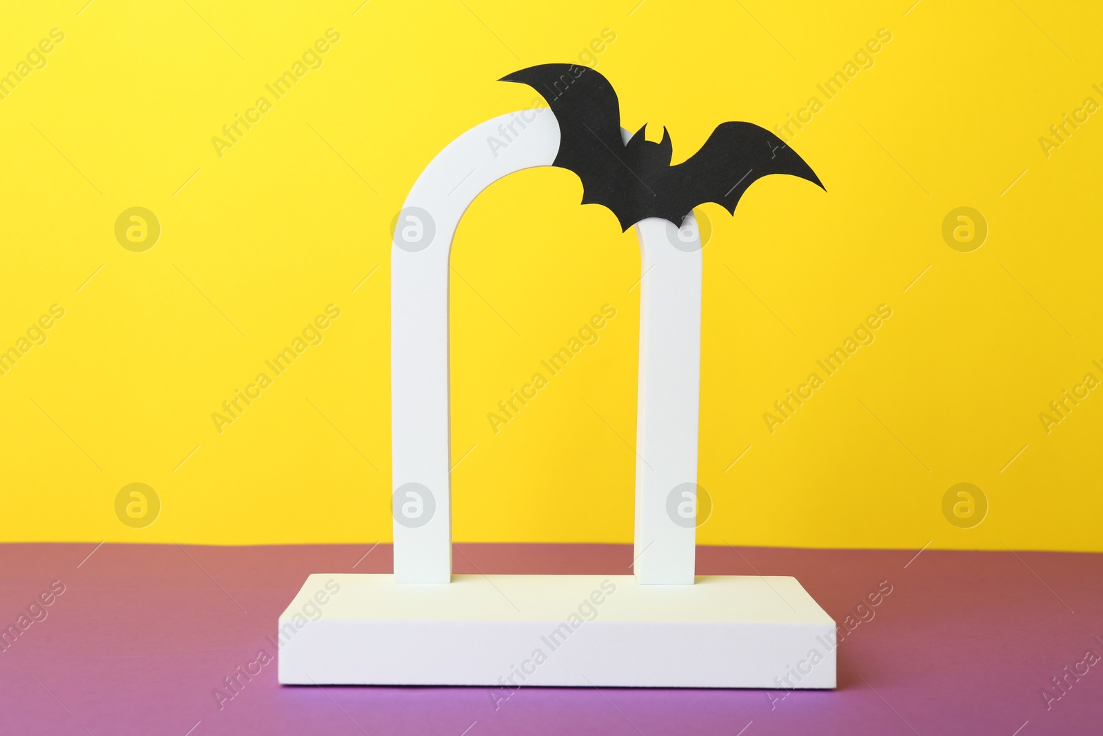 Photo of Halloween celebration. Podium and paper bat on purple table against yellow background. Space for text