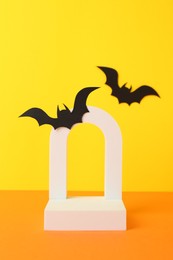 Halloween celebration. Podium and paper bats on orange table against yellow background, space for text