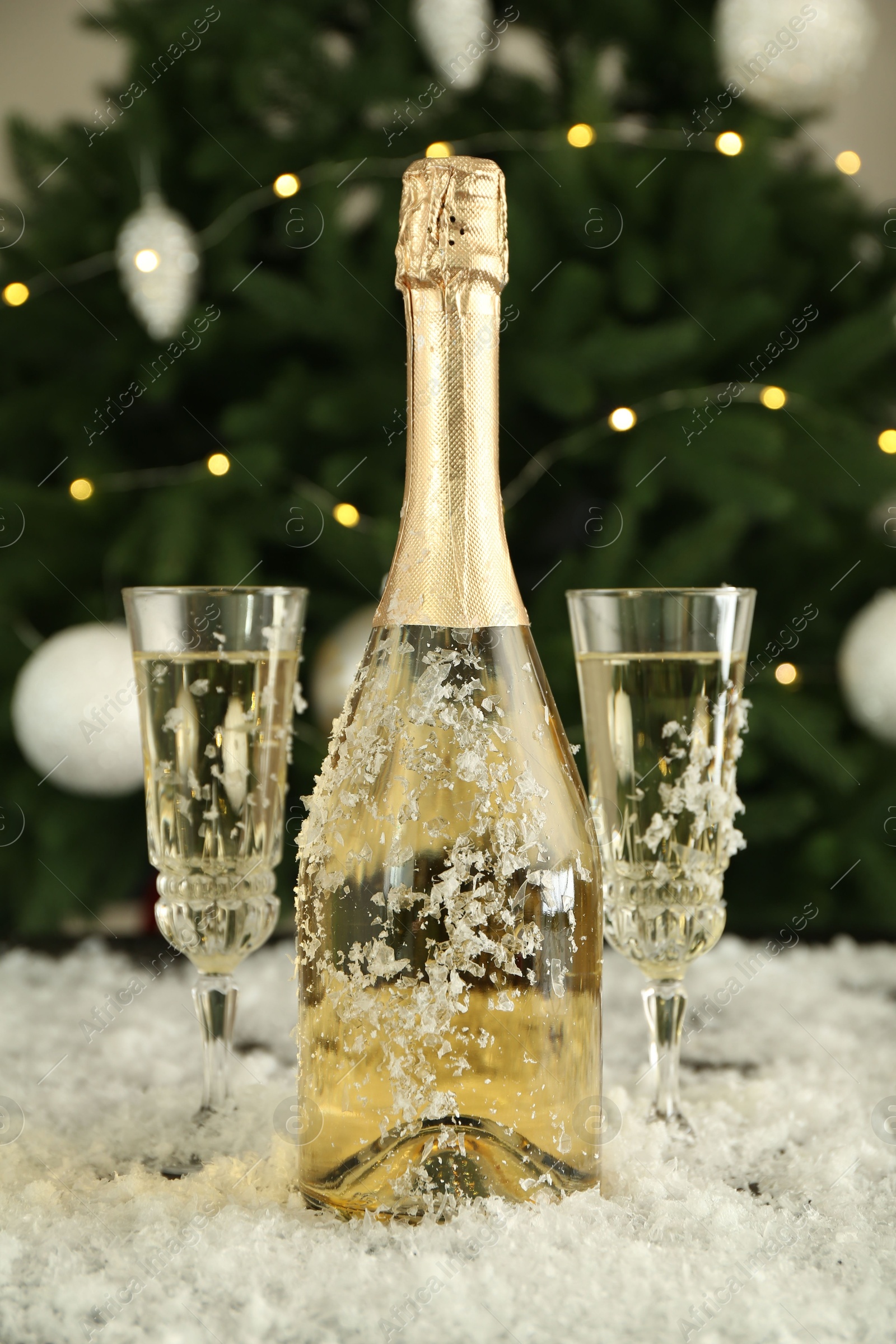 Photo of Sparkling wine in bottle, glasses and artificial snow on blurred background