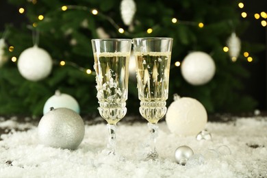 Photo of Sparkling wine in glasses, baubles and artificial snow on blurred background