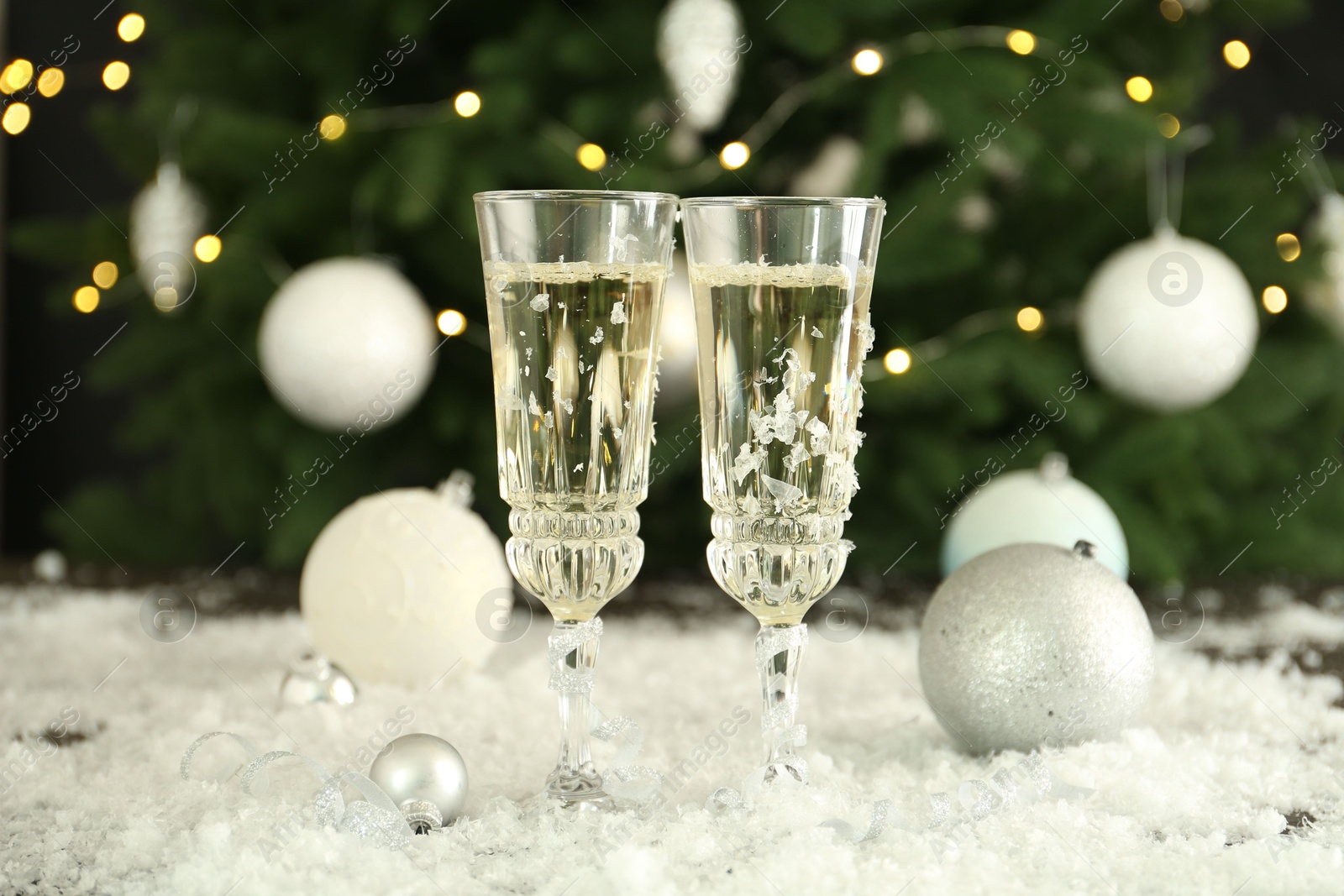 Photo of Sparkling wine in glasses, baubles and artificial snow on blurred background