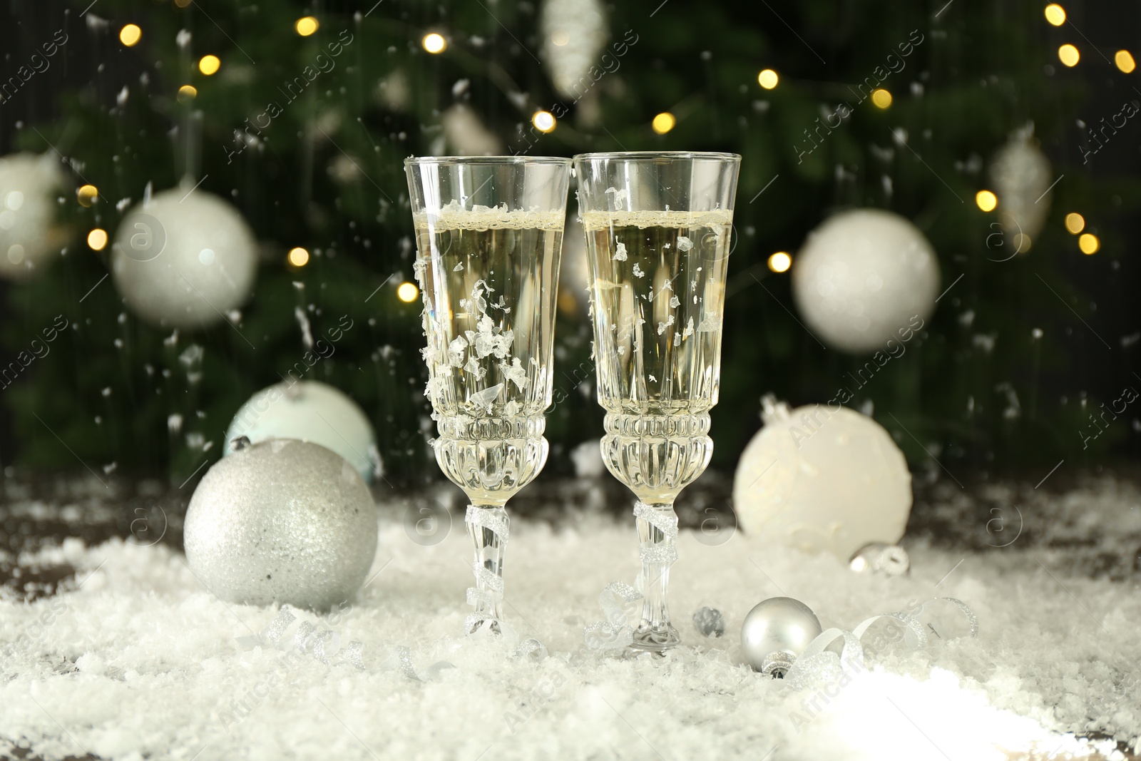 Photo of Sparkling wine in glasses, baubles and artificial snow on blurred background