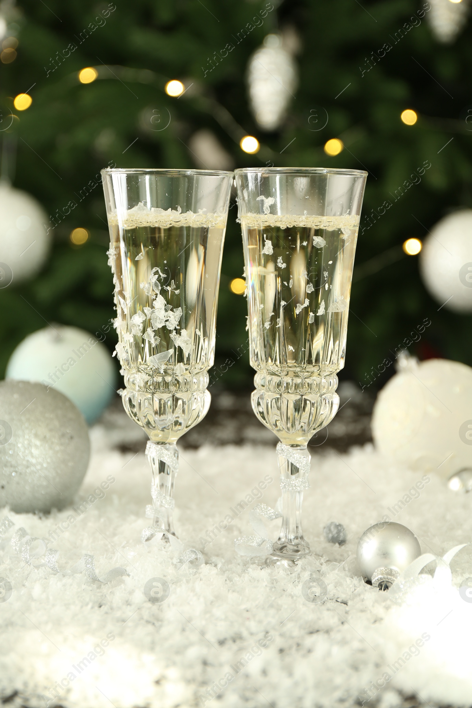 Photo of Sparkling wine in glasses, baubles and artificial snow on blurred background