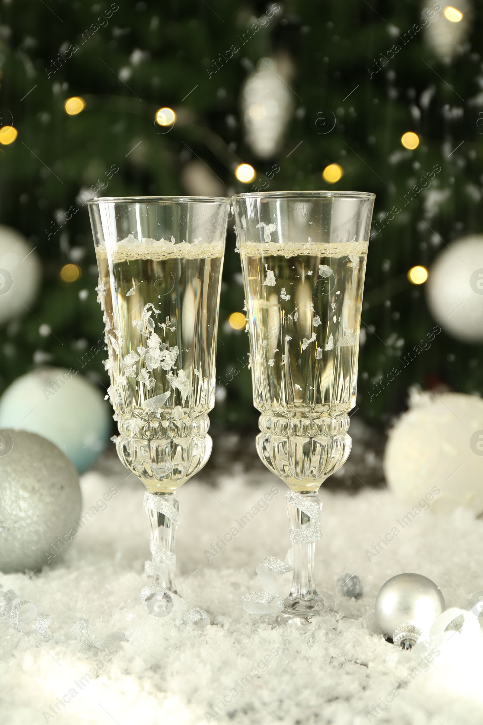 Photo of Sparkling wine in glasses, baubles and artificial snow on blurred background