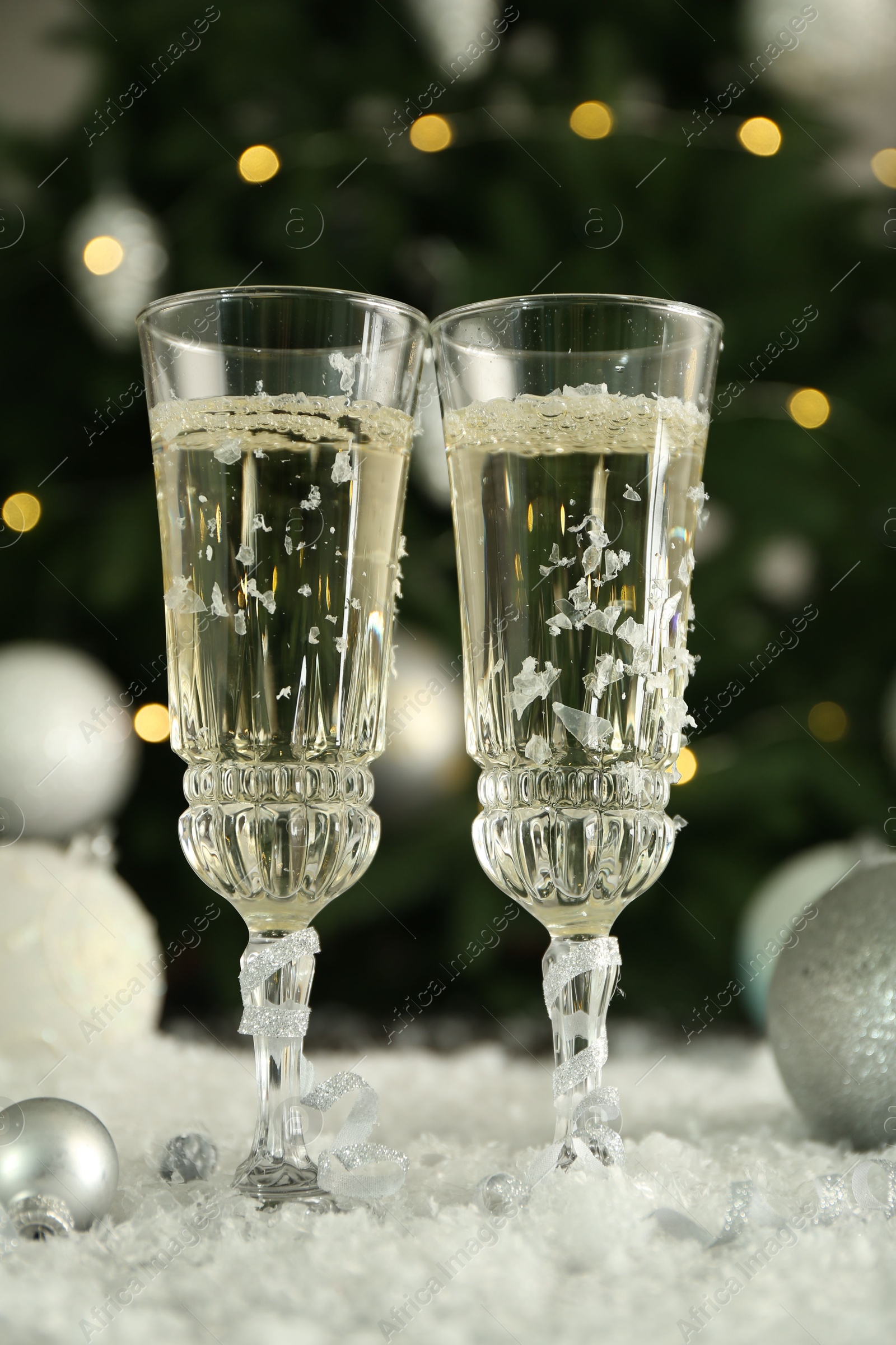 Photo of Sparkling wine in glasses, baubles and artificial snow on blurred background