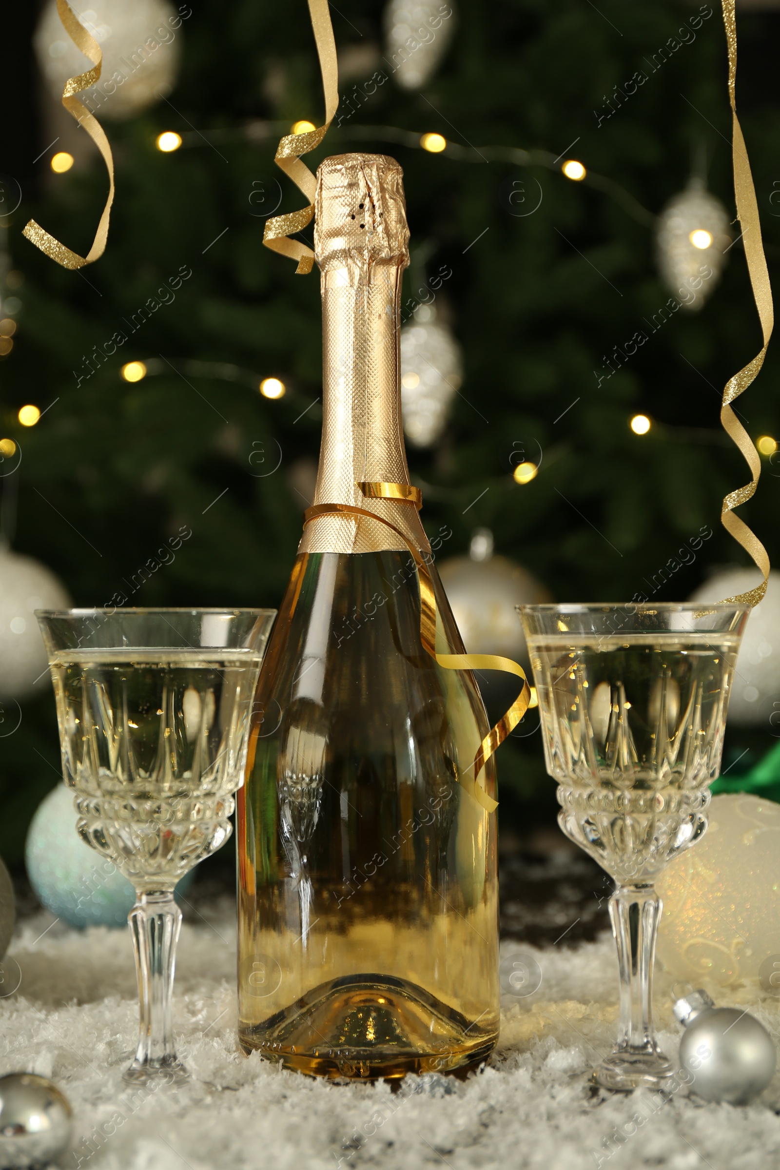 Photo of Sparkling wine in bottle, glasses, baubles and artificial snow on blurred background