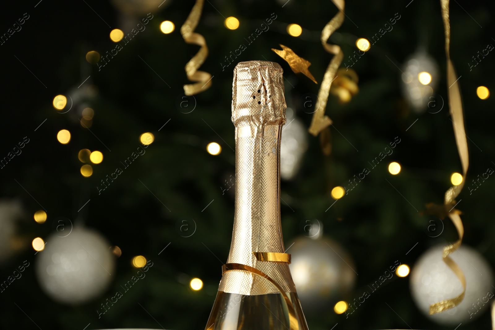 Photo of Bottle with sparkling wine and streamers against blurred lights, closeup