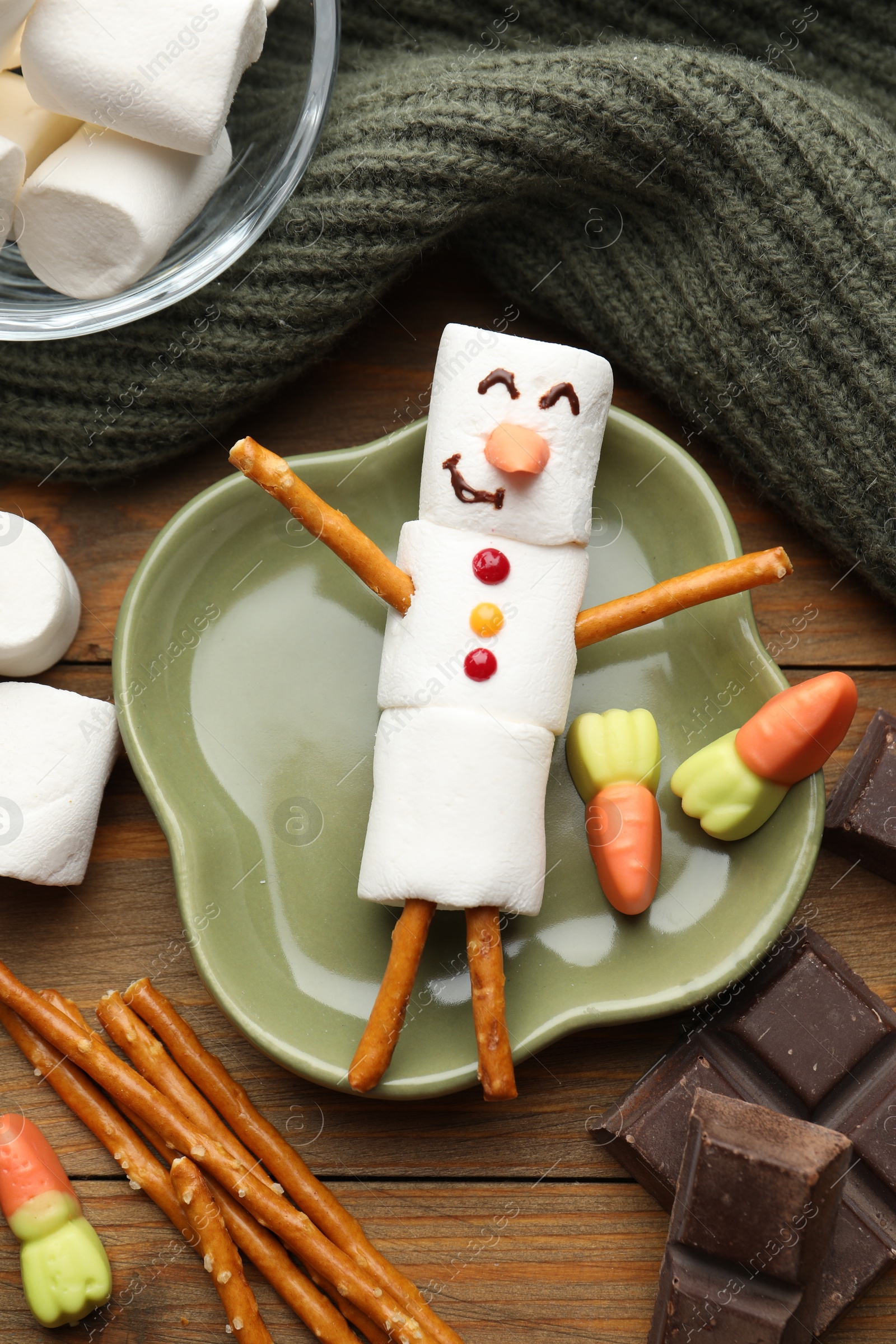 Photo of Funny snowman made with marshmallows, candies, chocolate and salty sticks on wooden table, flat lay