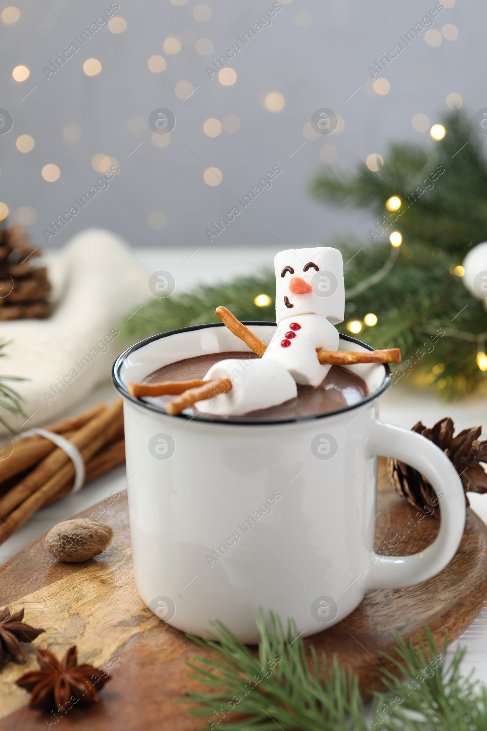 Photo of Funny snowman made with marshmallows in cup of hot chocolate and winter decor on white table against Christmas lights