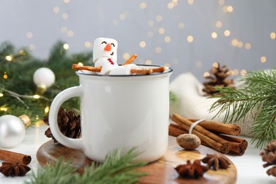 Photo of Funny snowman made with marshmallows in cup of hot chocolate and winter decor on white table against Christmas lights