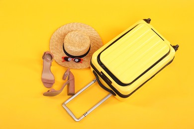 Photo of Suitcase with wicker hat, sunglasses and shoes on yellow background, above view