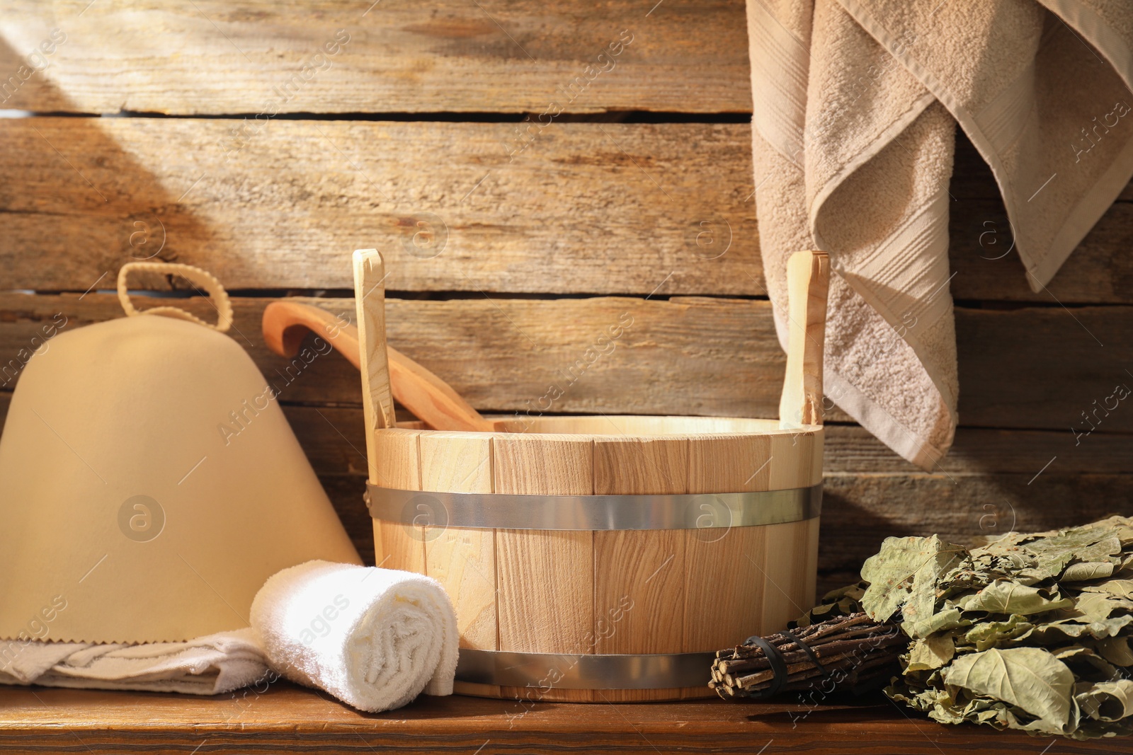 Photo of Sauna equipment. Bucket with ladle, felt wool hat, oak whisk and towels on wooden table indoors