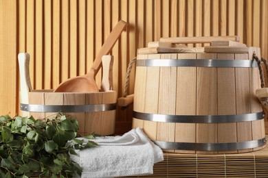 Photo of Sauna equipment. Buckets, birch whisk, ladle, and towel on bamboo bench near wooden wall