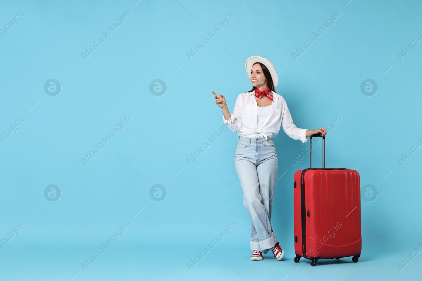 Photo of Happy young woman with suitcase pointing at something on light blue background, space for text