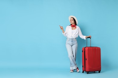 Happy young woman with suitcase pointing at something on light blue background, space for text