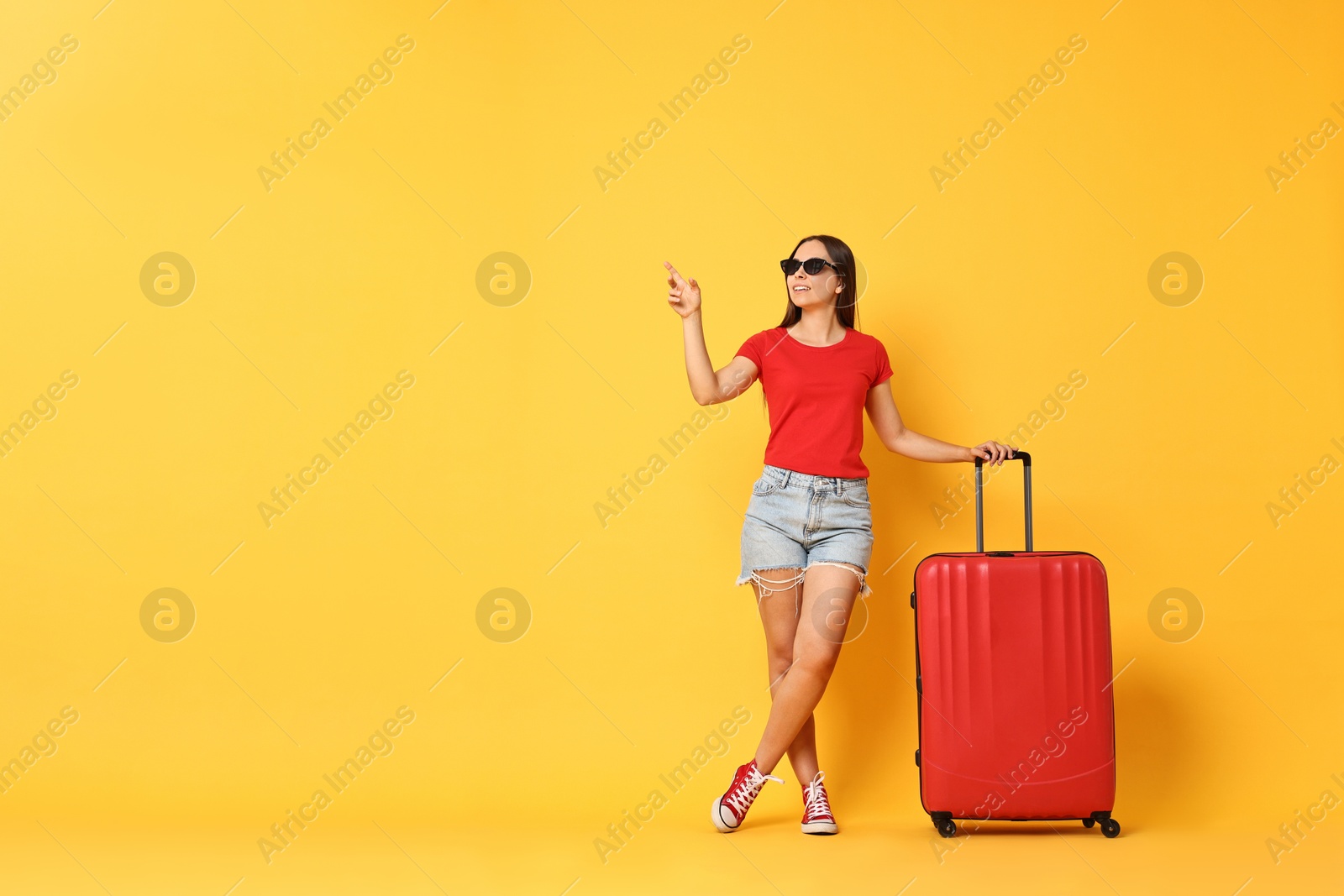 Photo of Happy young woman in sunglasses with suitcase pointing at something on orange background