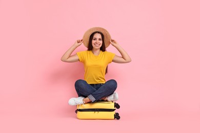 Happy young woman sitting on suitcase against pink background